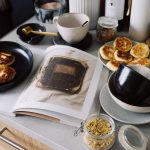 High angle of cookbook and cooked fritters on plates placed on table near utensils and ingredients in cozy kitchen at home