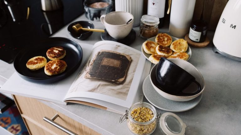 High angle of cookbook and cooked fritters on plates placed on table near utensils and ingredients in cozy kitchen at home