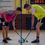 Two teenagers face off in a floorball match inside a sports gym with equipment visible in the background.