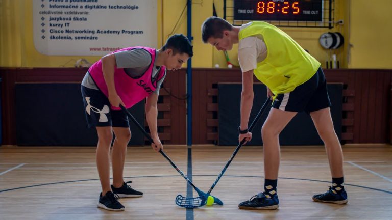 Two teenagers face off in a floorball match inside a sports gym with equipment visible in the background.